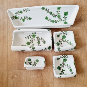 May include: A set of five white ceramic bathroom accessories. Each piece is decorated with green ivy vines and leaves. The set includes a rectangular tray, a soap dish, and three small square dishes.
