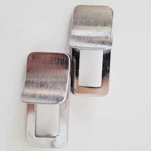 Op de afbeelding: Twee zilverkleurige, rechthoekige geldclips met een geborstelde metalen afwerking. Elke clip heeft een scharnierende bovenkant die opent en een witte binnenkant onthult. De clips zijn ontworpen om contant geld of kaarten vast te houden.