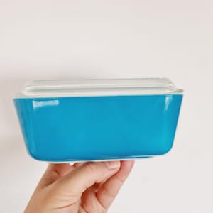 Peut inclure: Un plat à four rectangulaire Pyrex turquoise avec un couvercle en verre transparent. Le plat est tenu par une main sur fond blanc. Le plat a une finition brillante et un design rétro classique.