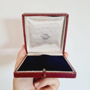 Puede incluir: Caja de joyería roja abierta con interior de satén blanco y base de terciopelo azul oscuro. La caja tiene un pequeño emblema plateado dentro de la tapa y un cierre dorado.