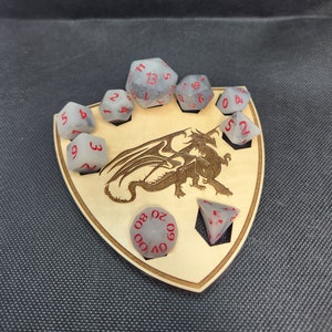 May include: A wooden dice tray in a shield shape, featuring a laser-etched dragon design. The tray holds several gray dice with red numbering. The dice are arranged around the dragon image, creating a thematic display for tabletop gaming.