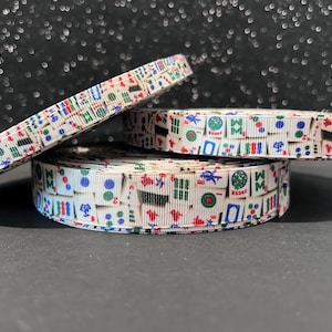 May include: Three rolls of white ribbon with a repeating pattern of colorful Mahjong tiles. The ribbon is stacked on top of each other, with one roll partially unraveled. The background is a dark, glittery surface.