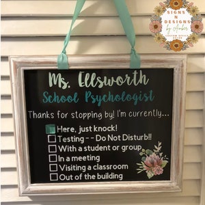 Könnte beinhalten: Ein gerahmtes Tafelschild mit einem weißen, verwitterten Rahmen und einem türkisfarbenen Band zum Aufhängen. Auf dem Schild steht "Ms. Ellsworth, Schulpsychologe" und enthält eine Checkliste für die Verfügbarkeit. Ein Blumendesign befindet sich in der unteren rechten Ecke.