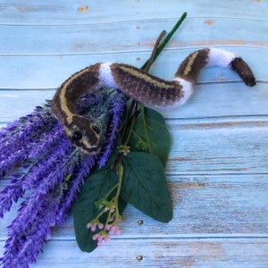 May include: A crocheted snake toy with brown and white stripes, curled around a bouquet of purple lavender and green leaves.