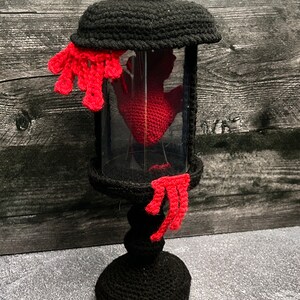 May include: A black crocheted lamp with a glass jar containing a red crocheted hand. The lamp has a red crocheted drip detail.