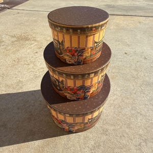 May include: Three round decorative hat boxes stacked on top of each other. The boxes have brown lids with a small dot pattern. The sides of the boxes have a cream and yellow striped pattern with a vintage illustration of a horse-drawn carriage.