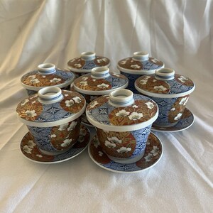 Puede incluir: Juego de siete tazas de té de estilo japonés con tapas y platillos. Las tazas y los platillos presentan un patrón geométrico azul y blanco con detalles florales y dorados. Las tapas tienen un pequeño asa.