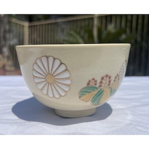 Puede incluir: Cuenco de cerámica color crema con diseños florales. El cuenco presenta un gran crisantemo blanco y dorado en un lado y un patrón floral más pequeño con detalles en verde y dorado en el otro. El cuenco tiene una base pequeña y circular.