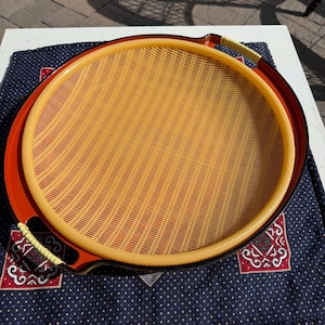 Puede incluir: Una bandeja redonda tejida en naranja y amarillo, con un borde rojo y dos asas. El centro de la bandeja tiene un patrón tejido, con un borde amarillo y asas envueltas en hilo amarillo y blanco. Descansa sobre una tela azul estampada.