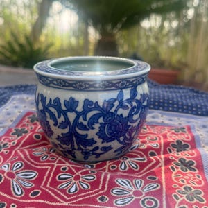 May include: A small, round ceramic planter featuring a white base with intricate blue floral and vine patterns. The rim is decorated with a repeating geometric border. The interior of the bowl is smooth and light blue, with a wide, open top.