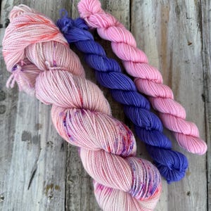 May include: Three skeins of yarn arranged side by side. The largest skein is a pale pink with vibrant purple and orange speckles. The middle skein is a solid deep purple, and the smallest skein is a solid light pink, all featuring a twisted texture.