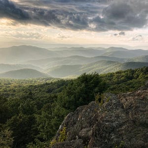 May include: A panoramic view of a mountain range with rolling hills covered in lush green trees. The sky is a mix of gray clouds and bright sunlight, creating a dramatic contrast.