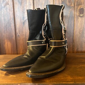 Könnte beinhalten: Ein Paar schwarze Lederstiefel mit spitzer Zehenpartie und wadenhohem Schaft. Die Stiefel verfügen über einen Riemen über dem Knöchel mit hellen Lederkanten, silberfarbene Metallbeschläge und vertikale Lederstreifen an der Seite.