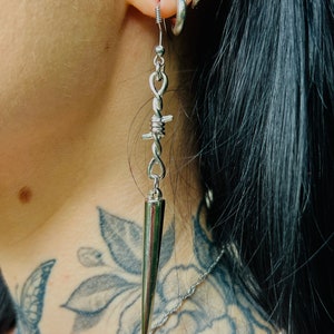 Puede incluir: Pendientes de plata con un diseño de alambre de púas y una larga punta puntiaguda. Los pendientes cuelgan de la oreja de una persona.