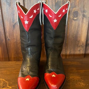 Könnte beinhalten: Ein Paar Cowboystiefel aus schwarzem und rotem Leder mit weißen Sternausschnitten an den roten Zehenkappen und den roten Schäften. Die Schäfte sind mit weißer Paspelierung und einem V-förmigen roten Kragen mit Stern- und Mondsichelmotiven verziert.
