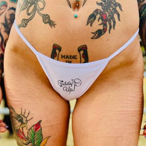 May include: White thong underwear with the text "Giddy Up!" and a cowboy hat graphic. The underwear has thin straps and is made of a soft material. The words "Made In" are also visible.
