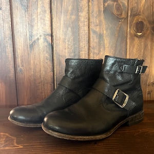 Könnte beinhalten: Ein Paar schwarze Lederstiefeletten mit abgerundeter Zehenpartie, niedrigem Blockabsatz und einem dekorativen Riemen über dem Spann, der mit einer silberfarbenen Metallschnalle befestigt ist. Ein weiterer Riemen mit Schnalle umschließt den Knöchel.