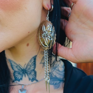 May include: Gold-toned oval earrings with a floral design and chain fringe. The earrings have a silver-colored chain and three spike charms. The earrings are hanging from a silver hoop. The earrings are part of a jewelry collection.