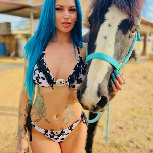 May include: A woman with blue hair and multiple piercings is wearing a cow print bikini and holding a black and white horse.