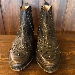 Könnte beinhalten: Ein Paar schwarze Stiefeletten mit einer strukturierten, reptilienartigen Musterung an der Schuhspitze und dem Vorderblatt. Die Stiefel sind mit zahlreichen kleinen, messingfarbenen Metallnieten am Obermaterial und an den Seiten verziert.
