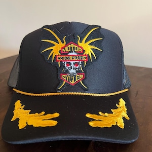 Könnte beinhalten: Schwarze Trucker-Cap mit Mesh-Rückseite und gebogenem Schirm. Auf der Vorderseite befindet sich ein Aufnäher mit den Worten "MOTOR CYCLES RIDE FREE" über einem Schädel- und Flügeldesign. Goldene Stickereien zieren den Schirm.