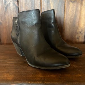 Könnte beinhalten: Ein Paar schwarze Lederstiefeletten mit abgerundeter Zehenpartie, einem Absatz in Holzschichtoptik und einem silberfarbenen Metallreißverschluss an der Außenseite jedes Schuhs. Die Stiefeletten haben eine glatte Oberfläche und eine strukturierte Form.