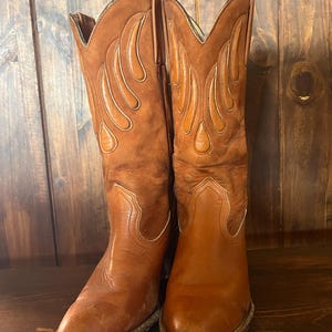 Könnte beinhalten: Ein Paar wadenhohe Cowboystiefel in einem mittleren Braunton. Die Stiefel haben einen Fuß aus glattem Leder und einen Schaft in Wildlederoptik mit dekorativen, flammenförmigen Lederapplikationen. Sie verfügen über eine spitze Zehenpartie.