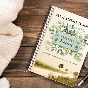 May include: A beige journal with a watercolor design of green leaves and a road leading to a field. The journal cover has the text "She is clothed in dignity" and "A Proverbs 31 Journal".