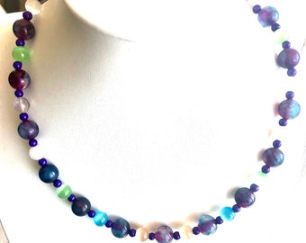 May include: A multi-coloured beaded necklace with purple, white and green beads. The necklace is made of round beads and is strung on a clear cord.