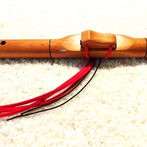 May include: A wooden Native American style flute with a natural finish. The flute has several finger holes and is adorned with red and black decorative ties. The instrument is designed for playing music.