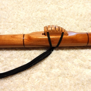 May include: A handcrafted wooden flute with a warm, reddish-brown finish. The flute features a carved wooden block with vertical grooves and black cord accents. The instrument is designed with three finger holes and a rounded end.