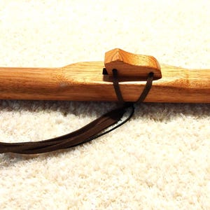 May include: A wooden Native American flute with a natural finish. The flute features a rectangular block and a leather strap. The instrument has three finger holes and a dark brown leather tassel. The flute is lying on a white, textured surface.