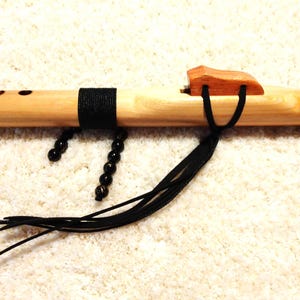 May include: A wooden Native American style flute with a natural finish. The flute has a wooden block, black accents, and decorative black beads. Black tassels hang from the wooden block. The flute is lying on a white, textured surface.
