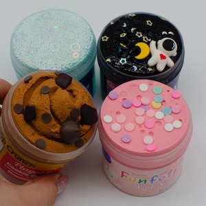 May include: Four containers of colorful slime. One is light blue with small beads, another is dark with an astronaut, a third is brown with chocolate chips, and the last is pink with confetti. The containers are clear with colorful lids.