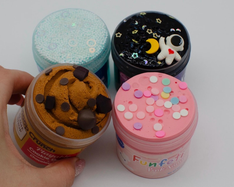 May include: Four containers of colorful slime. One is light blue with small beads, another is dark with an astronaut, a third is brown with chocolate chips, and the last is pink with confetti. The containers are clear with colorful lids.