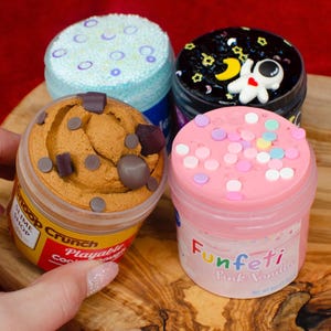 May include: Four jars of colorful slime. One jar has a cookie dough appearance with chocolate chip accents. Another has a pink top with sprinkles and the word "Funfetti". The other two jars have blue and black tops with decorative elements.