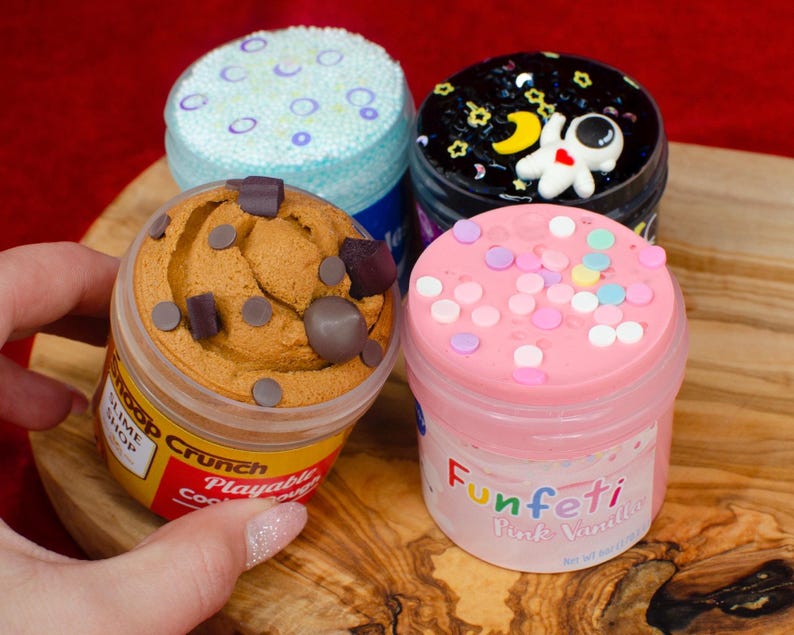 May include: Four jars of colorful slime. One jar has a cookie dough appearance with chocolate chip accents. Another has a pink top with sprinkles and the word "Funfetti". The other two jars have blue and black tops with decorative elements.