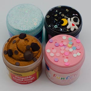 May include: Four jars of colorful slime. One jar has a light blue lid and is filled with small, round, white beads. Another has a black lid with a white astronaut and yellow moon. The third jar has a brown cookie dough design. The last jar has a pink lid with colorful sprinkles.