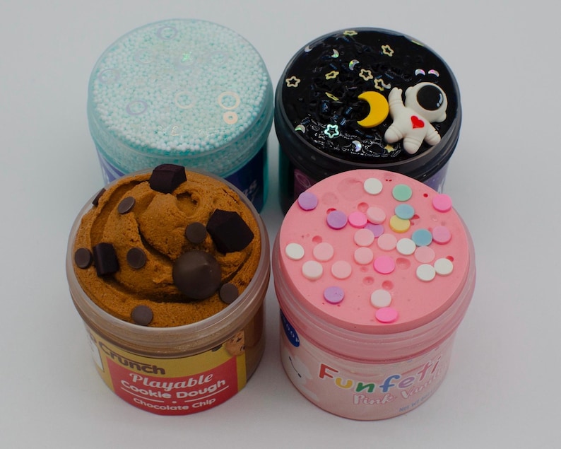 May include: Four jars of colorful slime. One jar has a light blue lid and is filled with small, round, white beads. Another has a black lid with a white astronaut and yellow moon. The third jar has a brown cookie dough design. The last jar has a pink lid with colorful sprinkles.
