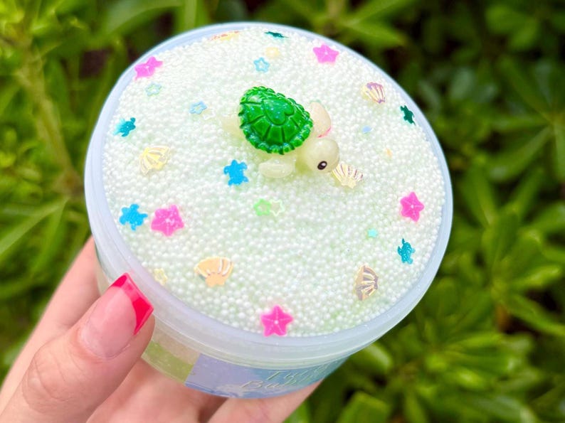 K&ouml;nnte beinhalten: Ein durchsichtiger Plastikbeh&auml;lter gef&uuml;llt mit einer hellgr&uuml;nen Substanz, verziert mit wei&szlig;en Schaumstoffb&auml;llchen, bunten stern- und muschelf&ouml;rmigen Streuseln und einer kleinen gr&uuml;nen Schildkr&ouml;tenfigur. Der Beh&auml;lter wird in einer Hand gehalten.