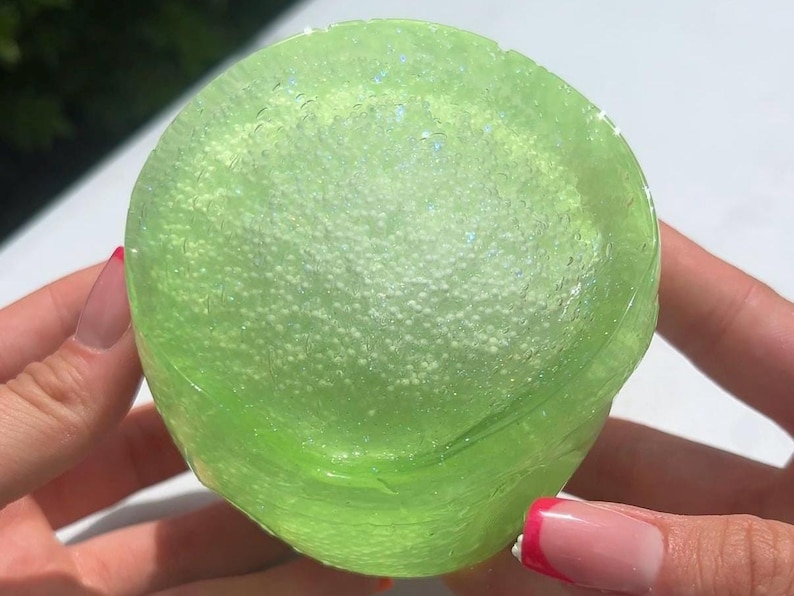 K&ouml;nnte beinhalten: Nahaufnahme eines durchscheinenden, limettengr&uuml;nen Schleimballs. Der Schleim ist mit kleinen, wei&szlig;en, kugelf&ouml;rmigen K&uuml;gelchen gef&uuml;llt. Der Schleim hat ein gl&auml;nzendes Aussehen und ist leicht irisierend. Das Bild ist gut beleuchtet und hebt die Textur und Farbe des Schleims hervor.