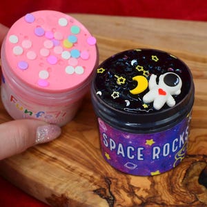 May include: Two small jars of slime on a wooden surface. One jar is pink with colorful confetti and the word "Funfetti". The other jar is black with an astronaut figure, stars, and the words "Space Rocks".