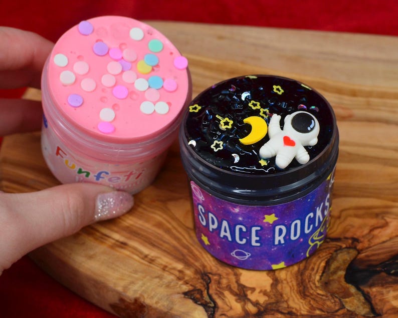 May include: Two small jars of slime on a wooden surface. One jar is pink with colorful confetti and the word "Funfetti". The other jar is black with an astronaut figure, stars, and the words "Space Rocks".