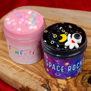 May include: Two jars of colorful slime sit on a wooden surface. One jar is pink with confetti and the text "Funfeti Pink Vanilla". The other jar is black with a small astronaut figure and the text "Space Rocks".