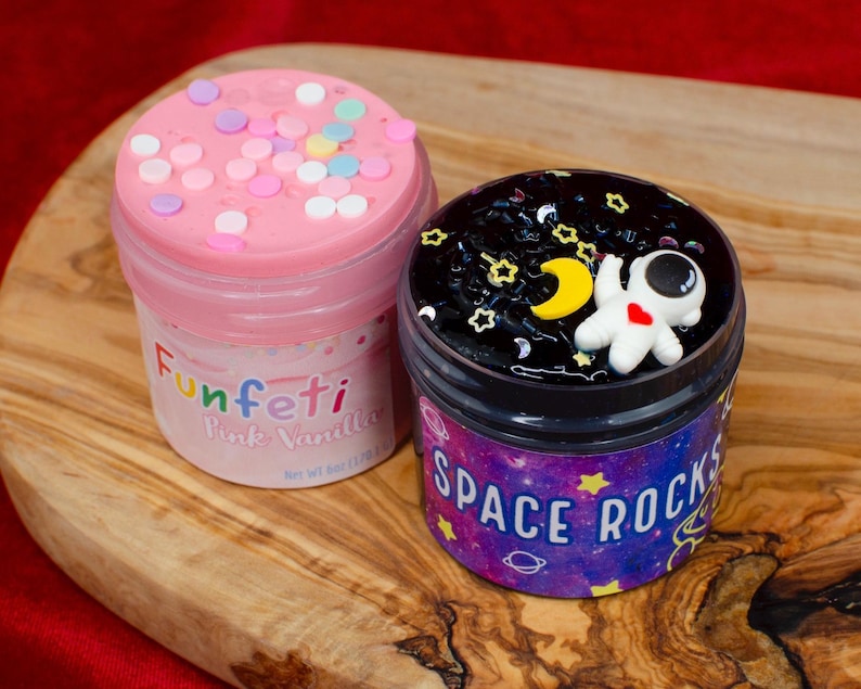 May include: Two jars of colorful slime sit on a wooden surface. One jar is pink with confetti and the text "Funfeti Pink Vanilla". The other jar is black with a small astronaut figure and the text "Space Rocks".