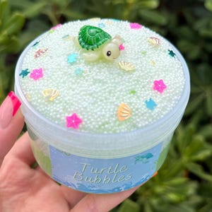 Puede incluir: Un recipiente de plástico transparente lleno de slime verde claro, decorado con pequeñas cuentas blancas, confeti colorido en forma de estrella y concha, y una pequeña figura de tortuga. El recipiente tiene la inscripción "Turtle Bubbles".