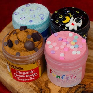 以下が含まれることがあります： カラフルで質感のあるプレイ・ドゥの4つの容器。1つはチョコレートチップ入りの茶色で、「Playable Cookie Dough Chocolate Chip」と表示されています。もう1つはスプリンクル入りのピンクで、「Funfetti Pink Vanilla」と表示されています。その他は青と黒の蓋が付いています。