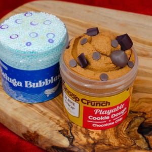 May include: Two containers of slime on a wooden surface. One is blue with the text "Beluga Bubbles" and a white whale illustration. The other is brown, resembling cookie dough, with chocolate chip accents and the text "Snoop Crunch Playable Cookie Dough Chocolate Chip".