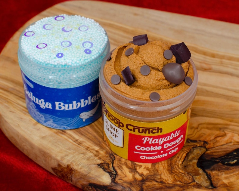 May include: Two containers of slime on a wooden surface. One is blue with the text "Beluga Bubbles" and a white whale illustration. The other is brown, resembling cookie dough, with chocolate chip accents and the text "Snoop Crunch Playable Cookie Dough Chocolate Chip".