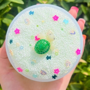 K&ouml;nnte beinhalten: Ein Beh&auml;lter mit Schleim mit hellgr&uuml;ner Basis, verziert mit kleinen wei&szlig;en Perlen und bunten Verzierungen. Die Dekorationen umfassen rosa Sterne, blaue Sterne, gr&uuml;ne Puzzleteile und Muschelformen. In der Mitte befindet sich eine kleine Schildkr&ouml;tenfigur.